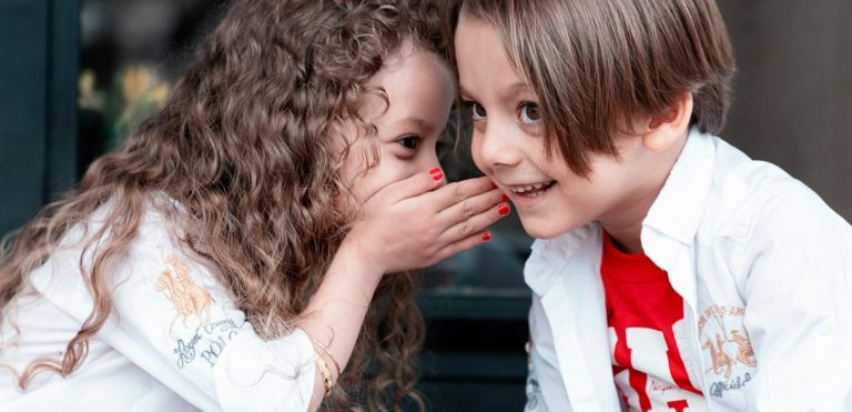 Sprachtherapie: Mädchen flüstert einem Jungen ins Ohr – Darstellung dafür, wie verbesserte Sprachkompetenz die Sprechfreude stärken kann.